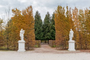 Alley in park with allegorical white stone sculptures against the hedges of trimmed deciduous trees with autumn leaves at overcast day clipart