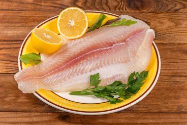 Defrosted fillet of the Pangasius, also known as basa fish on a big yellow dish with lemon halves and fresh herbs leaves on the old rustic table clipart