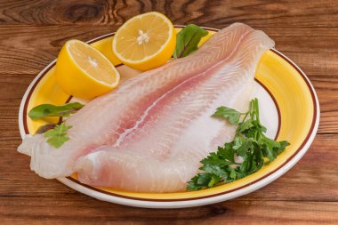 Pangasius 'un çözülmüş filetosu, aynı zamanda büyük sarı bir tabakta balık olarak da bilinir. Limon yarımları ve eski kırsal masada taze bitkiler, ön cephede yakın plan.