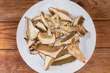 Dried slices of the brown caps and stems of Boletus edulis on a white dish on an old rustic table, top view clipart