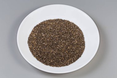 Soluk gri-kahverengi benekli chia tohumu yığını gri arka planda beyaz bir tabakta