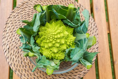 Kırpılmış saplı taze Romanesco brokoli 'nin başı ve ahşap bir masa üzerindeki cam kasede yapraklar.