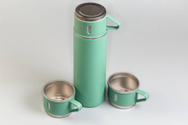 Closed small light green metal domestic vacuum flask covered with lid-cup and two same cups beside on a gray background clipart