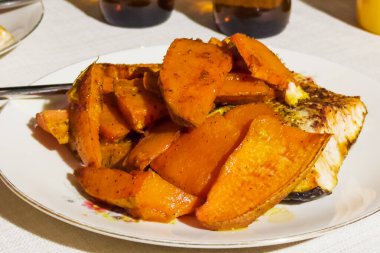 Portion of the slices of sweet potato tubers in their skin and fish piece baked in a oven on a dish on a table clipart