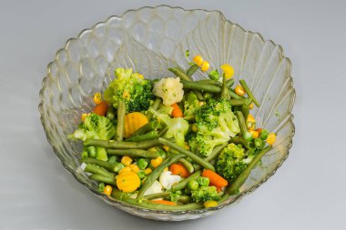 Farklı sebzelerin taze kaynatılmış karışımı gri arka plandaki cam salata kasesinde.