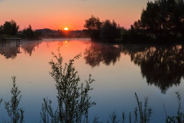 Sakin suyla dolu gölet ve ağaçlarla kaplanmış zıt kıyılar, suyun üzerinde sabah sisi ve uzak çayırlar, yaz gündoğumunda güneşin sudaki yükselişinin yansıması.