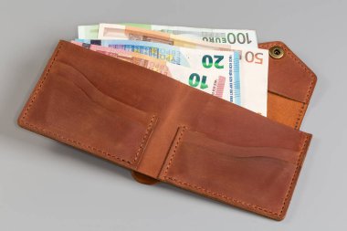 Klasik tarzda katlanmış kahverengi deri cüzdan. Gri arka planda birkaç farklı banknot var.