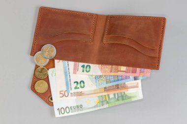 Klasik tarzda katlanmış kahverengi deri cüzdan. İçinde birkaç banknot ve bozuk para var. Gri arka planda üst görünüm.
