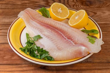 Pangasius 'un çözülmüş filetosu, büyük sarı bir tabakta balık olarak da bilinir, limon yarımları ve eski kırsal masada taze bitkiler, yakın plan.