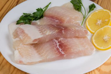 Pangasius 'un çözülmüş filetosunun çiğ parçaları. Limon dilimleri ve taze maydanozlu bir tabakta basa balığı olarak da bilinir.
