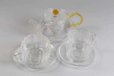 İçinde cam elek olan boş şeffaf bir çaydanlık ve iki farklı tutacağı olan iki farklı cam bardak ve gri arka planda iki adet boş duvar ve tabakları var.