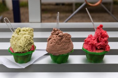 Üç porsiyon çikolatalı, fıstıklı ve ahudududulu dondurma. Açık bir kafede, yeşil, tek kullanımlık plastik bardaklarda küçük şeffaf kaşıklarla doldurulmuş.