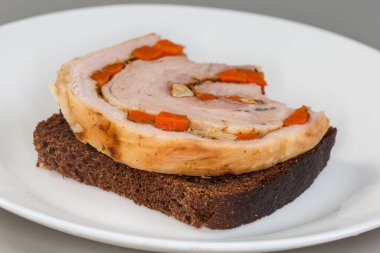 Kısmen açık sandviçi, fırında domuz eti rulosuyla, kahverengi ekmeğin üzerinde sebzelerle birlikte, bir tabağın üzerinde, ön planda seçici odak noktasında uzanıyor.