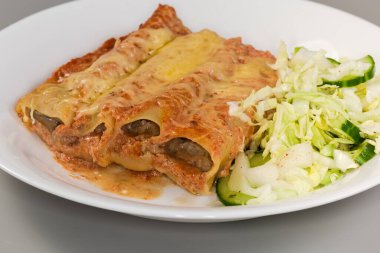 Fırınlanmış büyük silindirik makarna makarna cannelloni, peynirle doldurulmuş etle doldurulmuş, tabakta yeşil sebze salatası ile servis edilmiş, ön manzara seçici odak noktasında.