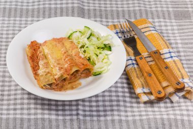 Masanın üzerine yeşil sebze salatası serpiştirilmiş, et dolmasıyla doldurulmuş büyük silindirik makarna tüpleri servis ediliyor.