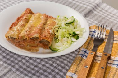 Fırınlanmış büyük silindirik makarna tüpleri, et dolgusu ile doldurulmuş, masaya yeşil sebze salatası serpiştirilmiş, yakın çekim