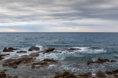 Sonbahar sabahında bulutlu gökyüzüne karşı, taştan bir sahil şeridindeki kayalar ve su yüzeyi arasında sörf dalgaları.