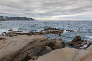 Kıyı kayaları ve deniz dalgaları arasında, bulutlu gökyüzüne karşı su yüzeyi ve sonbahar sabahında uzak kıyı tepeleri.