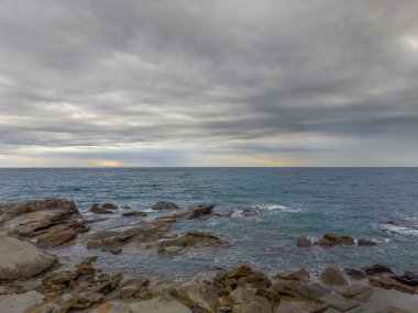 Sonbahar sabahında bulutlu gökyüzüne karşı, taştan bir sahil şeridindeki kayalar ve su yüzeyi arasında sörf dalgaları.