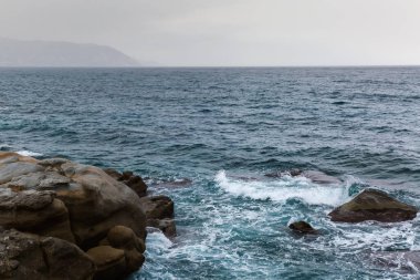 Kıyı kıyısında deniz yüzeyi, bulutlu gökyüzünün arka planında taştan kıyı şeridi ve bulutlu bir sonbaharda sisli kıyı tepeleri.