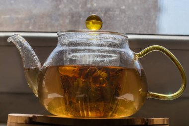 İçi cam elek ve sarı saplı cam şeffaf çaydanlıkta demlenmiş yeşil çay. Arka planda bulanık bir pencerenin ön görüntüsü.