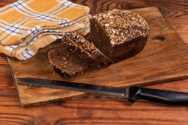 Kısmen dilimlenmiş kahverengi tahıl ekmeği, ahşap bir kesme tahtasına susam ve keten tohumu ile süslenmiş, ön planda tırtıklı mutfak bıçağı ile kırsal bir masada.