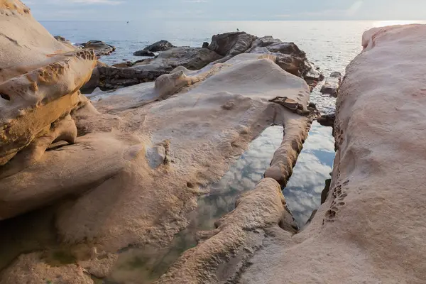 Güneşli bir sabahta, taştan bir sahil şeridindeki gri-kahverengi kumtaşı kayaları yıpranmış.