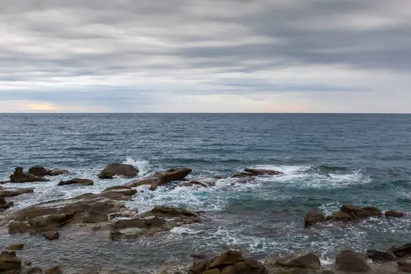 Sonbahar sabahında bulutlu gökyüzüne karşı, taştan bir sahil şeridindeki kayalar ve su yüzeyi arasında sörf dalgaları.