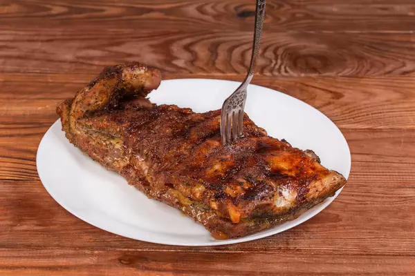 Baharatla pişirilmiş ve özel souse ile doldurulmuş domuz kaburgalarının tamamı, büyük beyaz bir tabağın çatalına saplanmış, kırsal bir masada, ön manzara seçici odak noktasında.