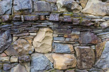 Doğal taşlarla inşa edilmiş ıslak, pürüzsüz istinat duvarının yüzeyi farklı formlar, renkler ve boyutlarda ve yağmurlu bulutlu bir günde kısmen yosun ile kaplanmış.