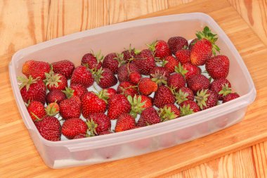 Yıkanmamış, organik yetiştirilmiş çilekler dikdörtgen düz plastik konteynırda kırsal bir masanın tahta kesme tahtasının üzerinde.