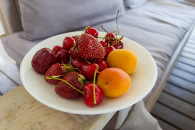 Taze çilekler, tatlı kirazlar ve kayısılar döşenmiş mobilyaların bulanık arka planında yakın plan beyaz bir tabakta.