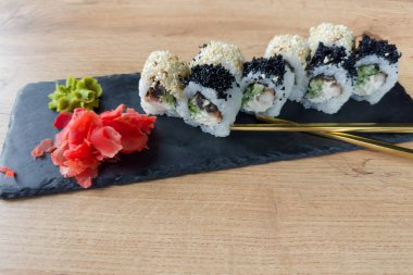 Sushi uramaki wasabi ve turşulu zencefil ile servis edilir siyah düz taş levha üzerinde metal çubuklar ile ahşap masa üzerinde, seçici odak