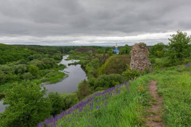 Ortaçağ taşından bir şatonun kalıntıları ile nehir vadisi ve yüksek sarp kıyılarda ahşap kilise ve bulutlu bir günde karşı kıyıda orman.