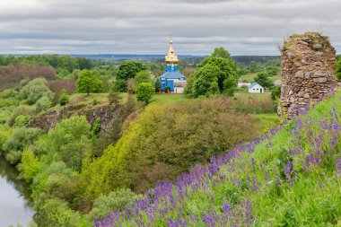 Dik nehir kıyısı, çiçek açan çayır adaçayıyla kaplanmış ve bulutlu bir günde, eski bir kale harabeleriyle ve ahşap kiliseyle kaplanmış.