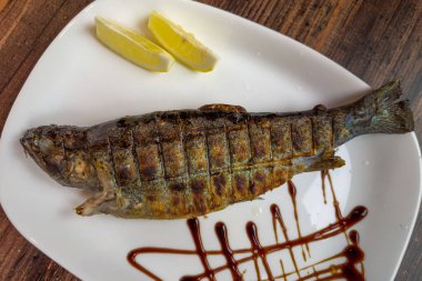 Nehir alabalığı ızgarada ızgarada kızartılıyor. Ahşap bir masada taze limon dilimleriyle servis edilen kömürün üstünde. Yakın plan.