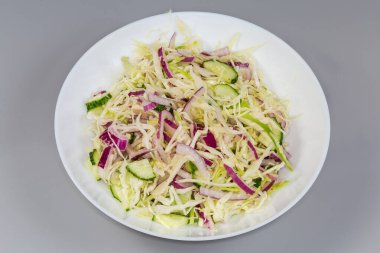 Dilimlenmiş taze beyaz lahanadan yapılmış sebze salatası, salatalık ve kırmızı soğan gri arka planda beyaz tabakta.