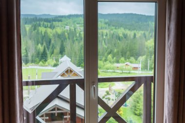 Window in mountain house with brown curtains along the edges and blurred mountain ridge view and neighboring buildings roofs on a foreground in spring overcast morning