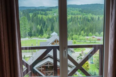 Forested mountain ridge, view through the window with brown curtains along the edges and roofs of neighboring buildings on a foreground in spring overcast morning