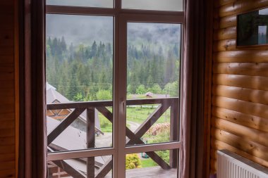 Dağ evindeki panoramik pencere ahşap panelli duvarlar, kenarları boyunca kahverengi perdeler ve bahar sabahında puslu dağ sırtının içerisinden manzara.