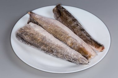 Arjantin hake 'sinin pişmemiş donmuş cesetleri mi, yoksa kafasız ve kuyruklu beyaz bir tabakta gri bir arka planda seçici bir odak noktasında yakın plan mı?