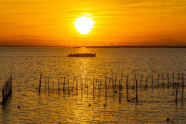 Turistlerin ve balıkçıların olduğu göl kenarında balık ağları ve güneşli bir yol olan tekne turuncu bir günbatımının arka planına karşı.