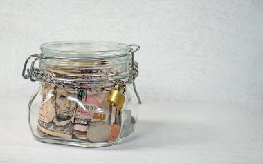 Gold padlock and chain secured around money jar on textured light gray background