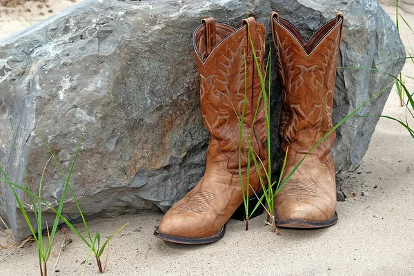 棕色的西式靴子 被一块大岩石和绿叶的草覆盖在沙滩上 — 图库照片
