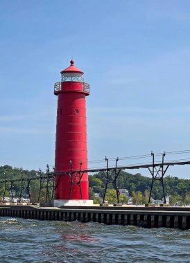 Canlı kırmızı bir Grand Haven Michigan deniz feneri açık mavi bir gökyüzüne karşı dimdik duruyor, yemyeşil ve sakin bir su önplanı var.
