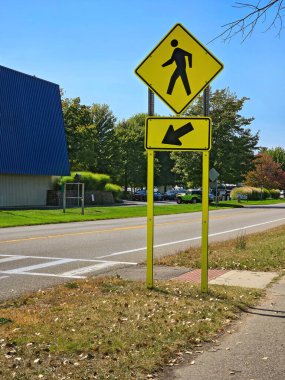 Üzerinde ok olan neon sarısı yaya geçidi işareti ağaçlık bir yolun yanında ve boş bir yolun yanında mavi çatılı bir bina gösteriliyor.