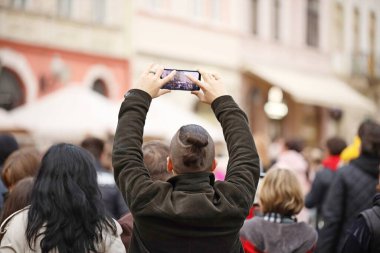 Şık bir saç kesimi olan genç bir adam cep telefonuyla kalabalığın arasında kollarını kaldırarak fotoğraf çekiyor. Konserde bir hayran, en sevdiği grubun performansının videosunu cihazında çekiyor.