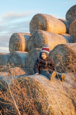 A cute little boy in autumn clothes and a knitted hat poses against a background of golden hay bales. Vertical photo.