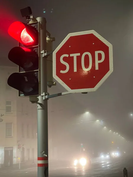 Vertical shot of stop sign next to traffic lights glowing red in foggy weather at night
