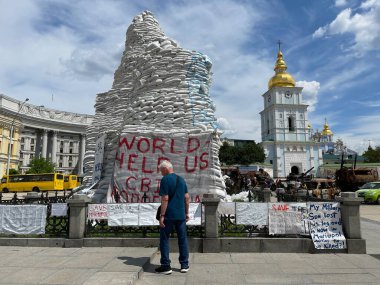 Ukrayna 'da savaş. 17 Haziran 2022 'de Ukrayna' nın Kyiv kentindeki müstahkem anıtlardan sallanan beyaz kumaşla yazılmış Dünya Yardım Derneği mesajının önünde duran kıdemli adam.